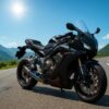 マットブラックに赤ラインが入ったカスタムCBR650Rが、青空の下で山道に停車しているシーン。カスタムマフラーやフェンダーレス仕様が映え、昼の陽光に包まれた雄大な山々と曲がりくねる道路が広がる16:9構図の高品質画像。