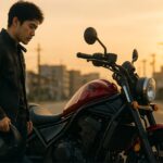 夕暮れの街中で赤いホンダ・レブル1100の横に立ち、購入を考えるようにバイクを見つめる日本人ライダー。
