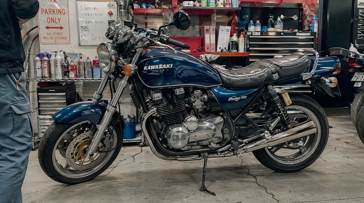 整備工場で点検・整備を受けるカワサキ・ゼファー750。工具やケミカル類が並ぶ作業場で、購入前の状態確認やメンテナンスの重要性が伝わる車体。