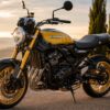 夕暮れの黄金色の光の中、上品な屋外の舗装路に停車したカワサキZ900RS風のバイクを、左向きのフロント斜めから写したリアルなヒーロー画像。丸型ヘッドライト、しずく型タンク、黒と黄色のクラシックなカラーリング、金色のホイール、直列4気筒エンジン、磨かれた金属パーツが強調され、背景には柔らかくぼけた山並みと樹木が見える。