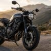 ゴールデンアワーの山道に停車した、ヤマハXSR700風のレトロモダンな黒いバイクを低い正面斜めアングルで捉えた写真風ビジュアル。背景には曲がりくねった山岳道路と夕日に照らされた山並みが広がり、バイクが主役としてシャープに写っている。