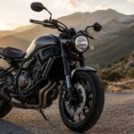 ゴールデンアワーの山道に停車した、ヤマハXSR700風のレトロモダンな黒いバイクを低い正面斜めアングルで捉えた写真風ビジュアル。背景には曲がりくねった山岳道路と夕日に照らされた山並みが広がり、バイクが主役としてシャープに写っている。
